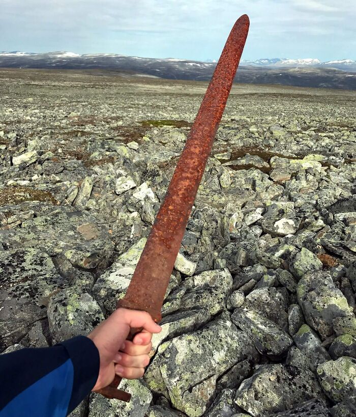 Hand holding a rusty ancient sword over rocky terrain, showcasing a bizarre historical artifact from a remote location.