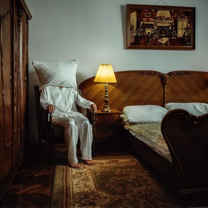 Surreal vintage photographic style showing a person with a pillow over their head in a dimly lit intimate bedroom setting.