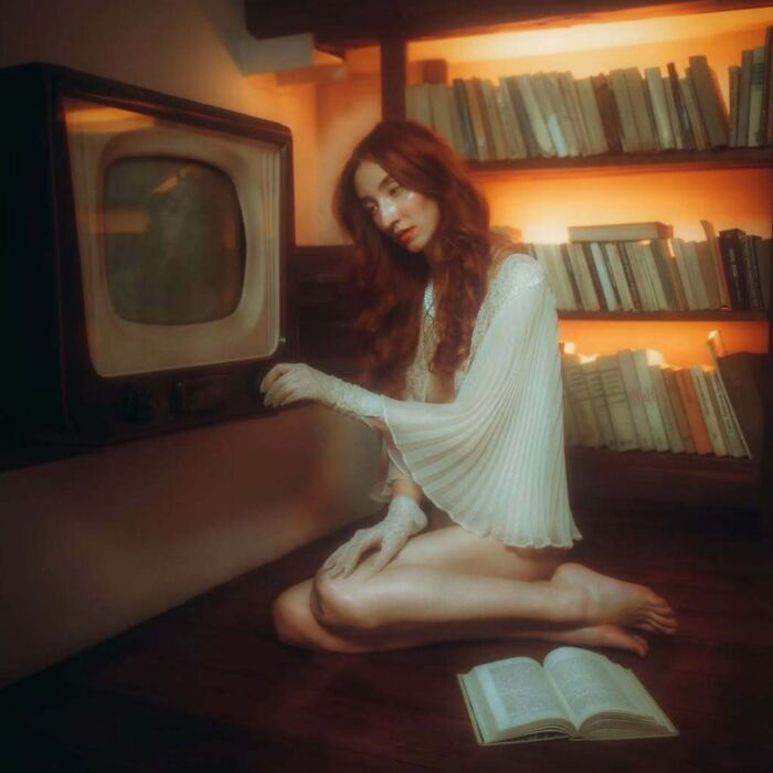 Woman in vintage style outfit kneeling by an old television in a softly lit room, captured with vintage cameras.