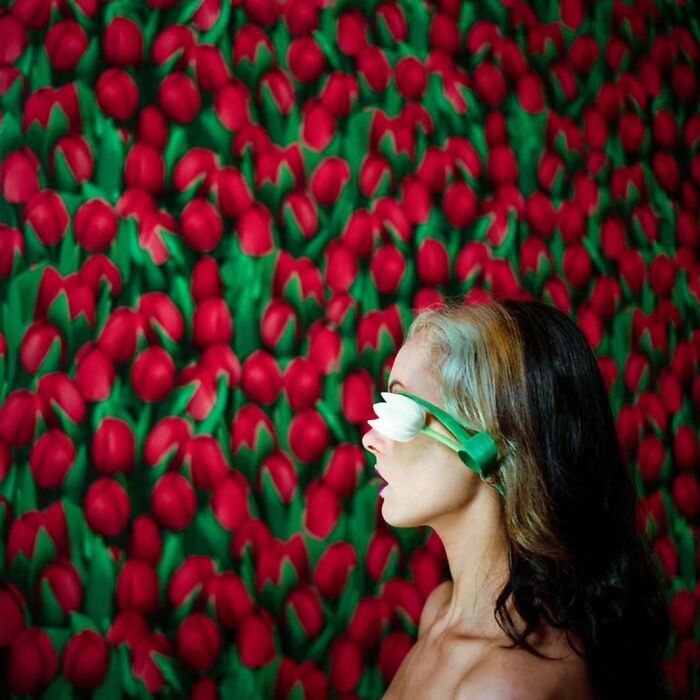 Woman with dark hair wearing a white flower blindfold, posing in front of a vibrant backdrop of red and green tulips, vintage camera style.