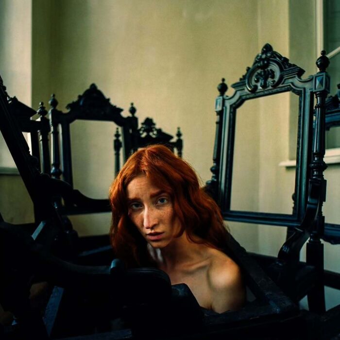 Woman with red hair in dim light posing among vintage chairs, captured in intimate cinematic-looking photography style.