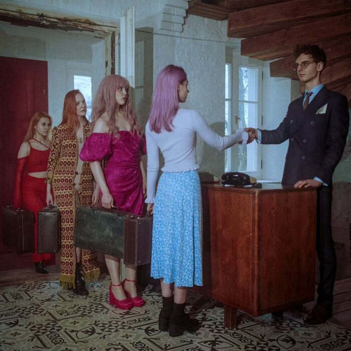 Vintage cameras capture intimate, cinematic-looking scene with four women holding suitcases and a man shaking hands indoors.