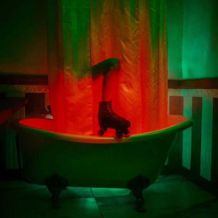 A vintage camera photo shows a leg with a roller skate emerging from a green-lit bathtub with a red shower curtain.