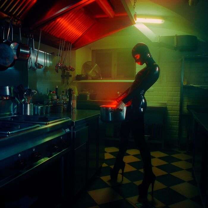 Person dressed in black latex suit standing in a dimly lit kitchen with vintage cinematic-looking lighting