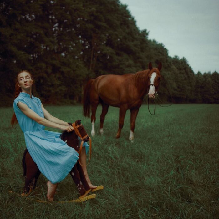 Woman in a blue dress poses on a rocking horse in a field with a real horse, captured with vintage cameras for cinematic shots.