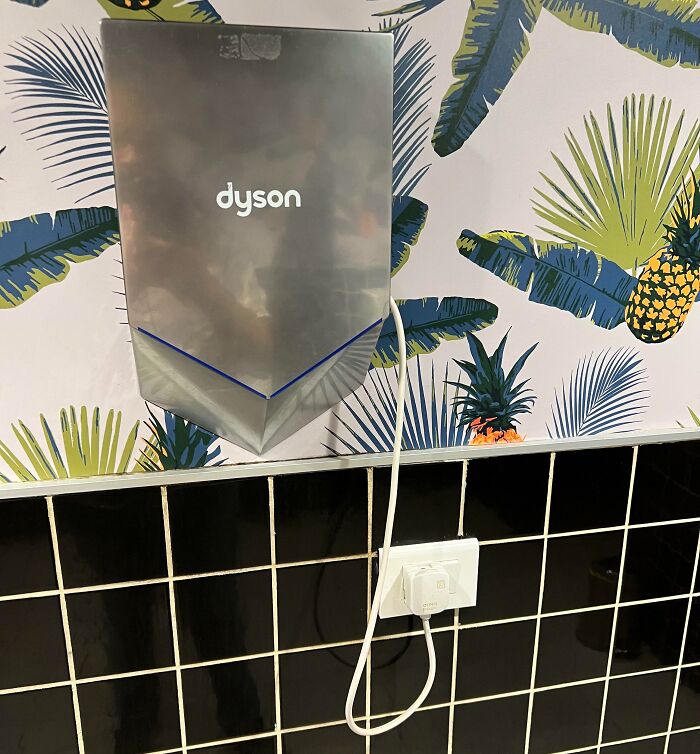 Dyson hand dryer with an exposed electrical cord plugged into a wall outlet on tiled wall with tropical wallpaper.