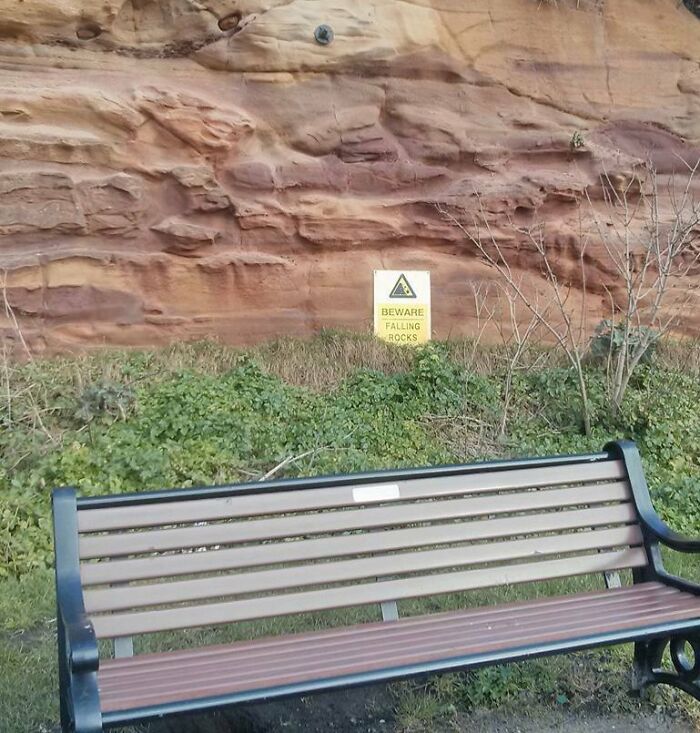 Park bench placed directly under a cliff with a dangerous warning sign about falling rocks in a risky design death trap scenario.