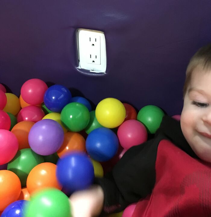 Child playing in colorful ball pit near a dangerous electrical outlet design posing death trap risk.