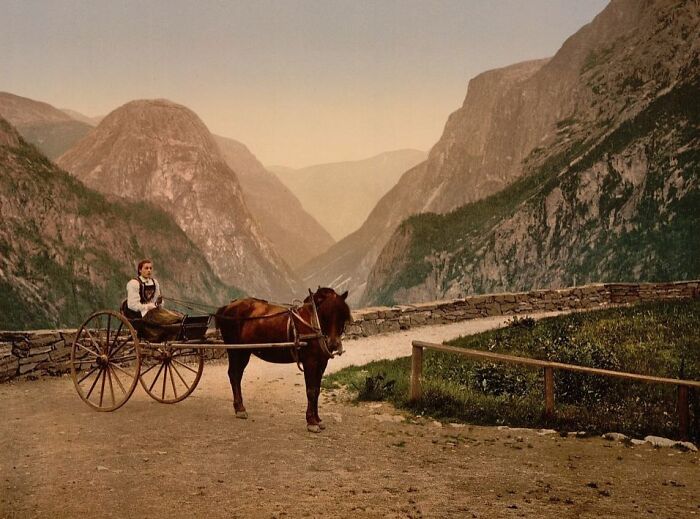 Colorized photo of a person in traditional clothing riding a horse-drawn cart in a mountainous cultural landscape.