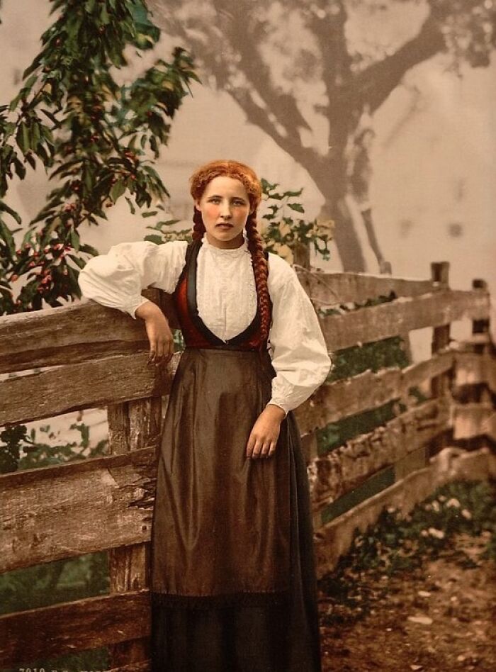 Colorized photo of a young woman in traditional dress leaning on a wooden fence revealing vibrant cultural past.