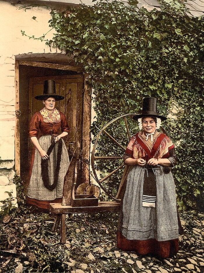 Two women in traditional dress with black hats beside a spinning wheel in a colorized photo of vibrant cultural past.