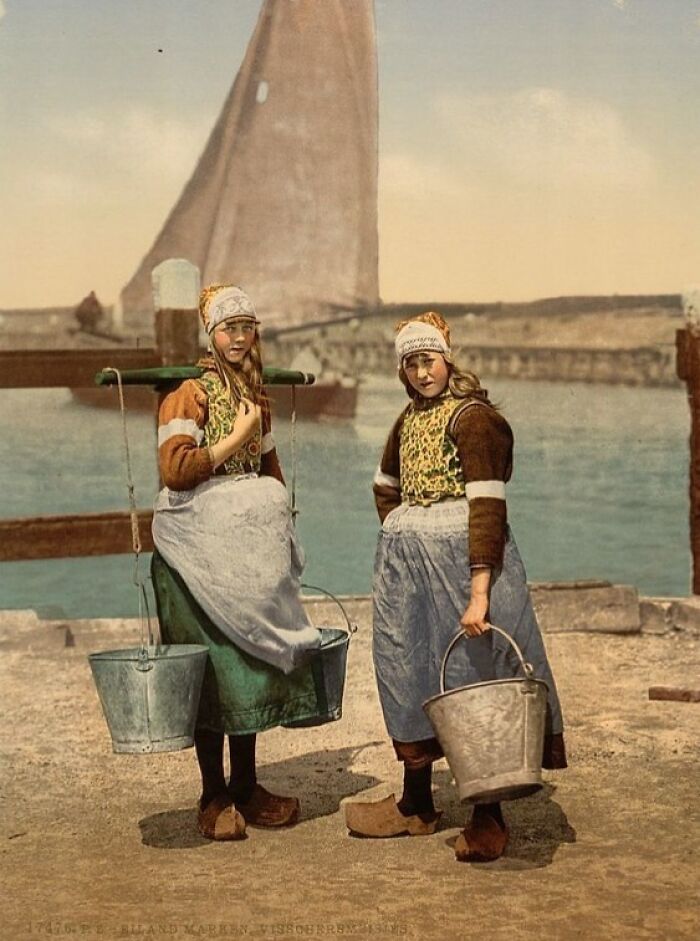 Two women in traditional clothing carrying buckets by the water, a colorized photo revealing vibrant cultural past.
