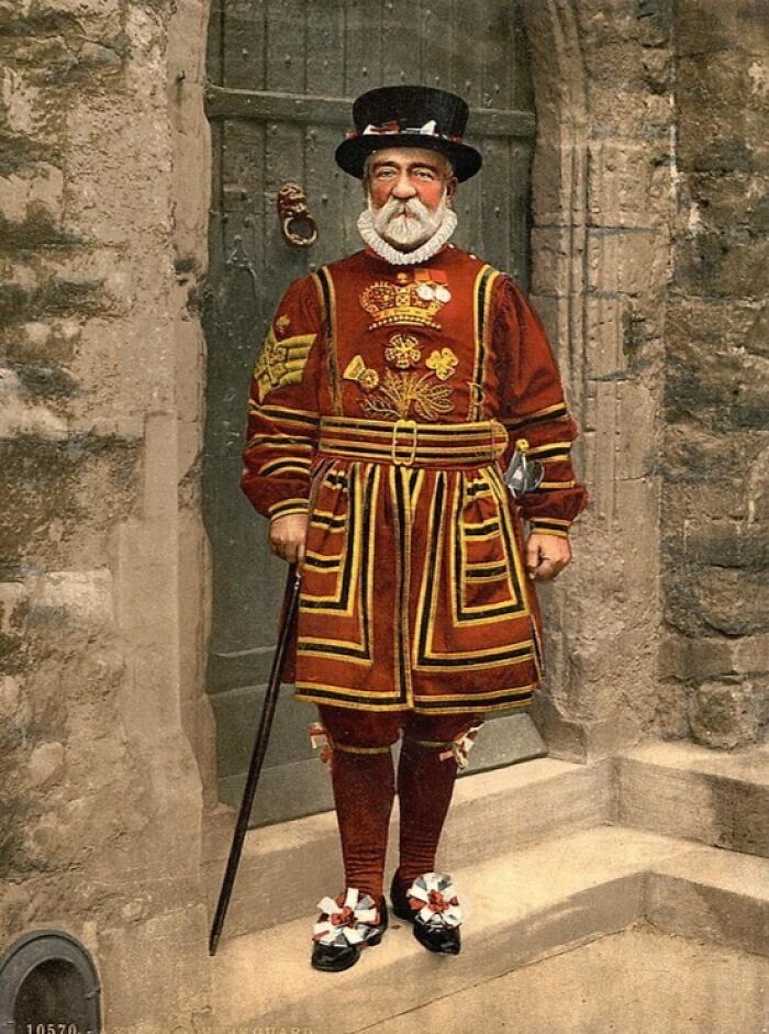 Colorized photo of a man in traditional vibrant cultural attire standing by a stone doorway showcasing historical dress.