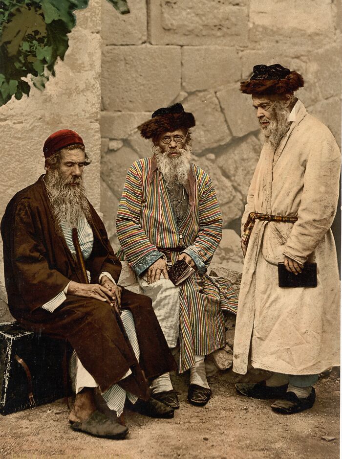 Three elderly men in traditional attire sitting and standing against a stone wall in a colorized cultural photo.