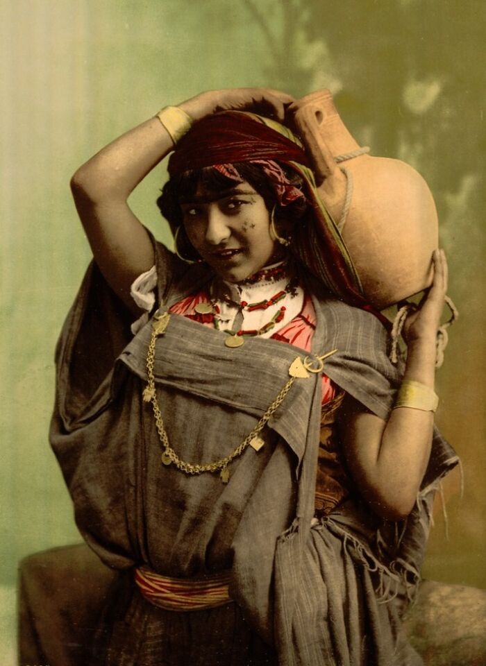 Young woman in traditional attire carrying a clay pot, colorized photo revealing vibrant cultural past.