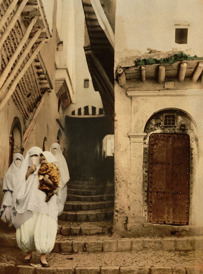 Women in traditional dress walking through a historic alleyway, showcasing vibrant cultural past in colorized photo.
