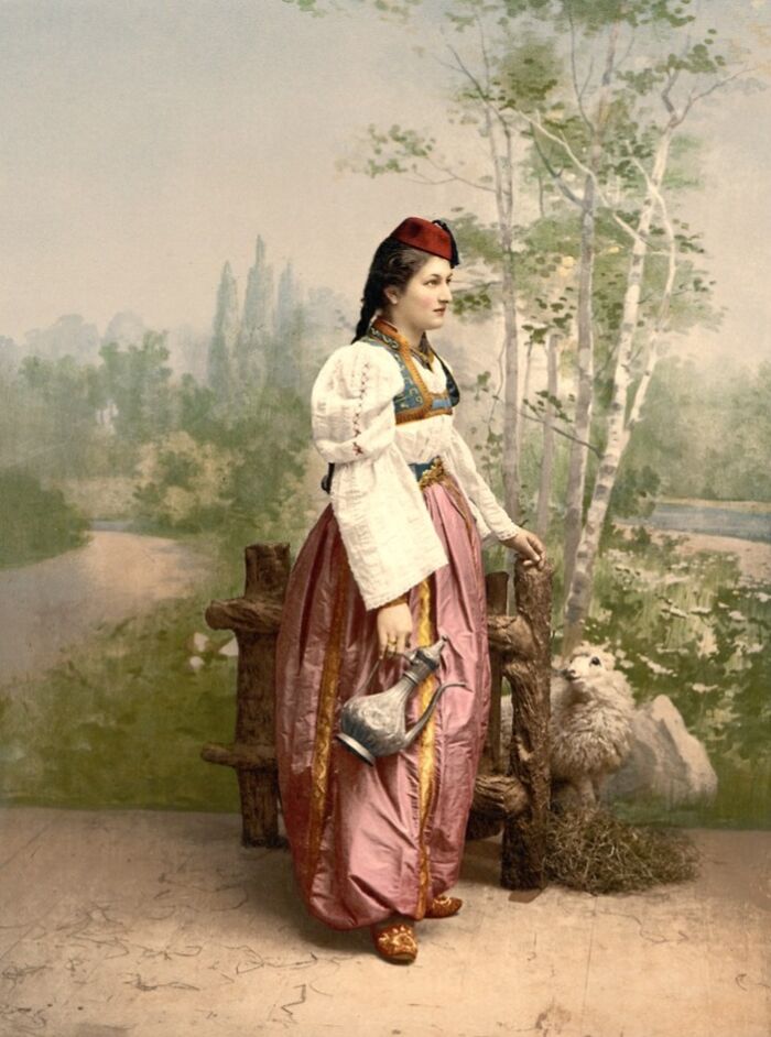 Woman in traditional dress holding a pitcher, standing by a rustic fence in a colorized photo revealing cultural past.
