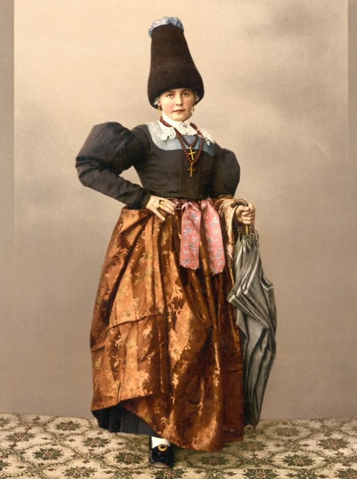 Young woman in traditional dress and tall hat holding an umbrella, showcasing vibrant cultural past in colorized photo.