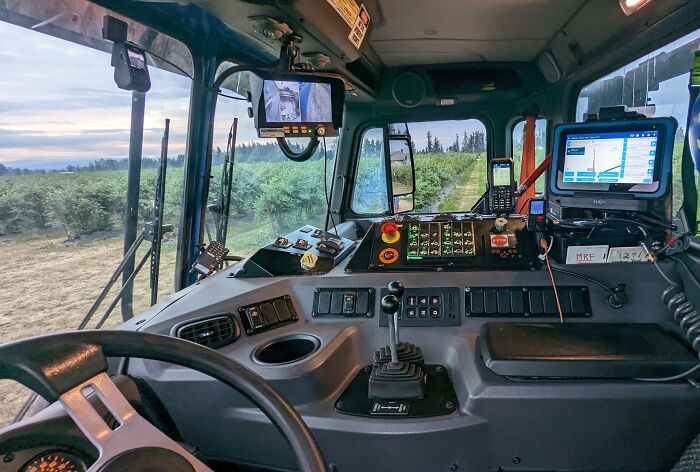 Cabin view of a high-tech agricultural vehicle with controls and screens driving through green fields, curious minds exploring technology.