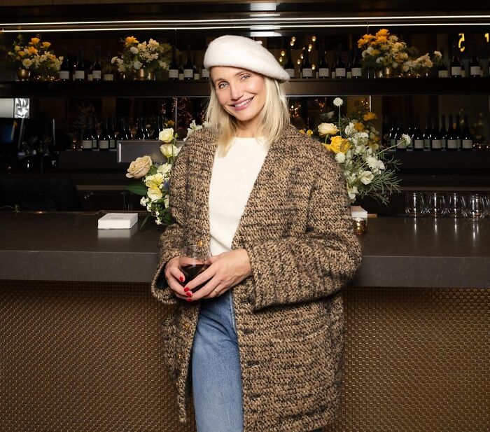 Blonde woman in a beret and cozy jacket holding a drink at a bar with flowers, relating to celebrity hygiene habits.