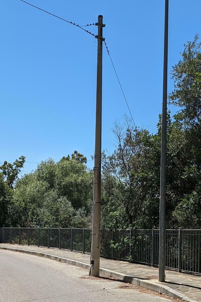 Streetlight pole with exposed wires posing a dangerous design hazard beside a sidewalk and metal fence under clear sky.