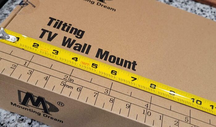 Measuring a tilting TV wall mount box with a yellow tape measure highlighting dangerous design risks.