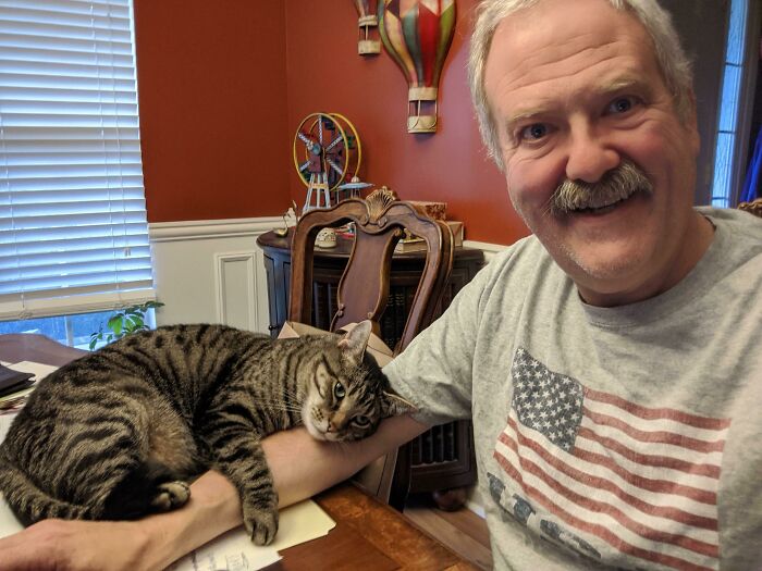 Man smiling at camera while a tabby cat rests on his arm at a wooden table in a home office setting, funny work-from-home meme concept.