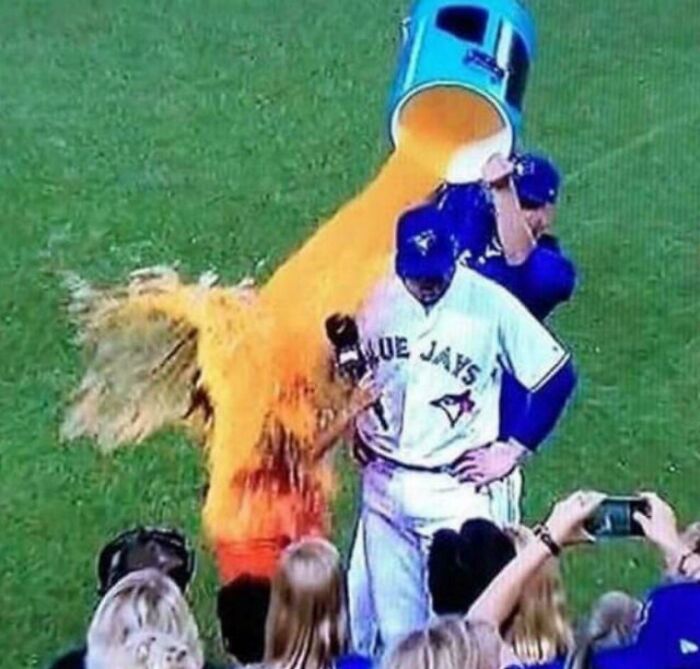 Baseball player getting drenched in orange liquid during a hilarious main character moment at a sports event.