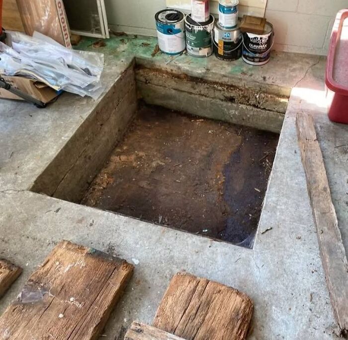 Concrete floor with a hidden pit inside a home, surrounded by paint cans and wooden planks in a forgotten space.