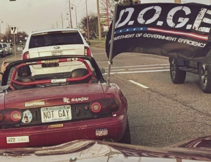 Red convertible car with a NOT GAY license plate and a large D.O.G.E flag on a city street, showing outrageous hot takes.