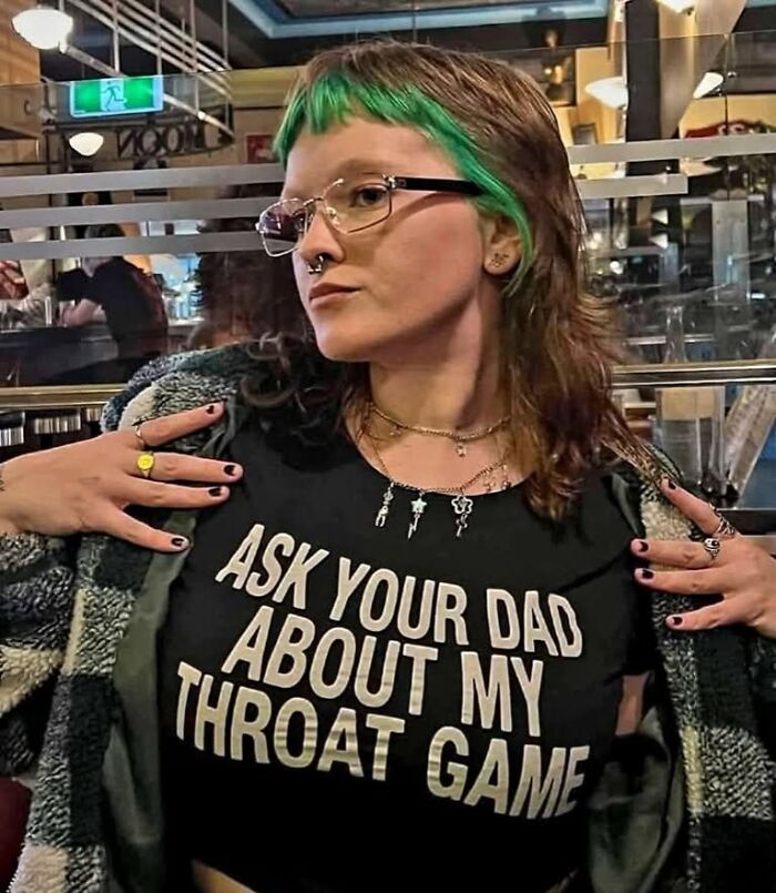 Person with green-streaked hair wearing glasses and a black shirt with text, showcasing outrageous hot takes in a casual indoor setting.
