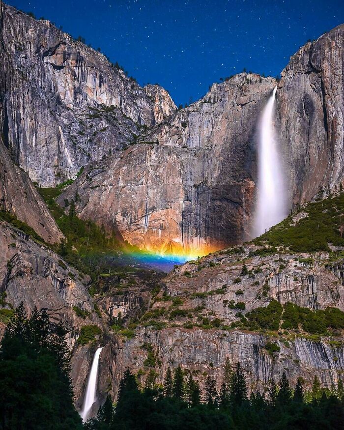 Waterfall with vibrant rainbow glowing at the base, set against rocky cliffs under a starry sky, capturing rare natural phenomena.