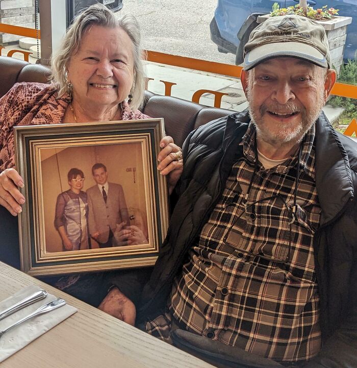Elderly couple smiling at a restaurant holding a framed photo of their younger selves showing love unchanged by time.