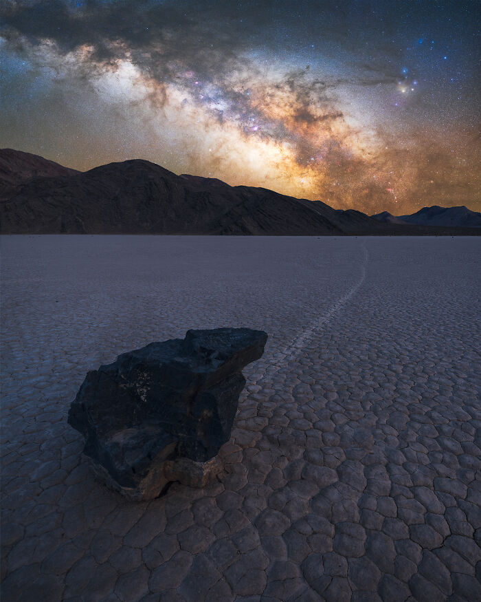 Dark rock on c*****d desert floor with Milky Way galaxy above, capturing rare natural phenomena under a starry night sky.