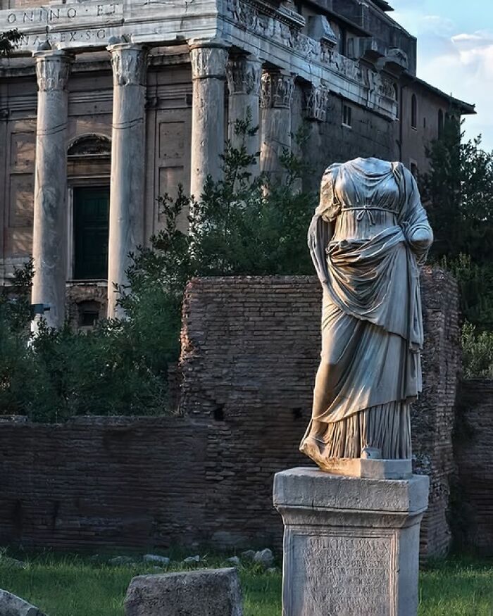 Ancient headless marble statue in front of classical archeological ruins surrounded by greenery and old brick walls.