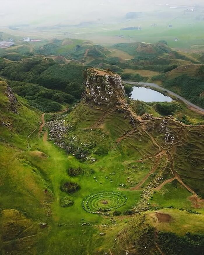 Aerial view of a green landscape featuring an ancient archeological site with circular stone formations and rugged hills.