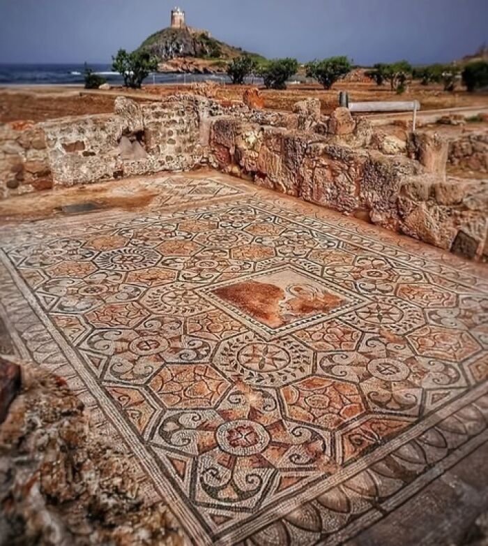 Ancient mosaic floor amid archeological ruins with a coastal landscape and lighthouse in the background.