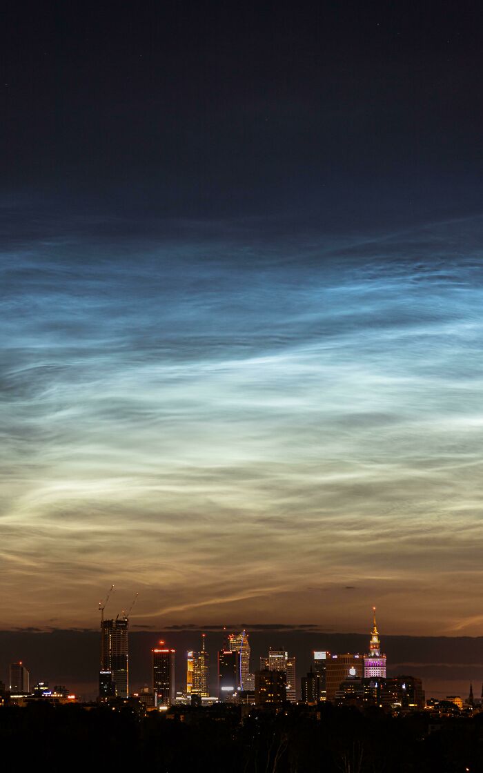 Nighttime city skyline under glowing noctilucent clouds, capturing rare natural phenomena with blue and white hues.