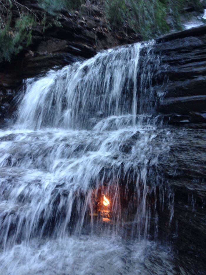 A rare natural phenomena showing a small eternal flame burning behind a cascading waterfall over layered rocks.