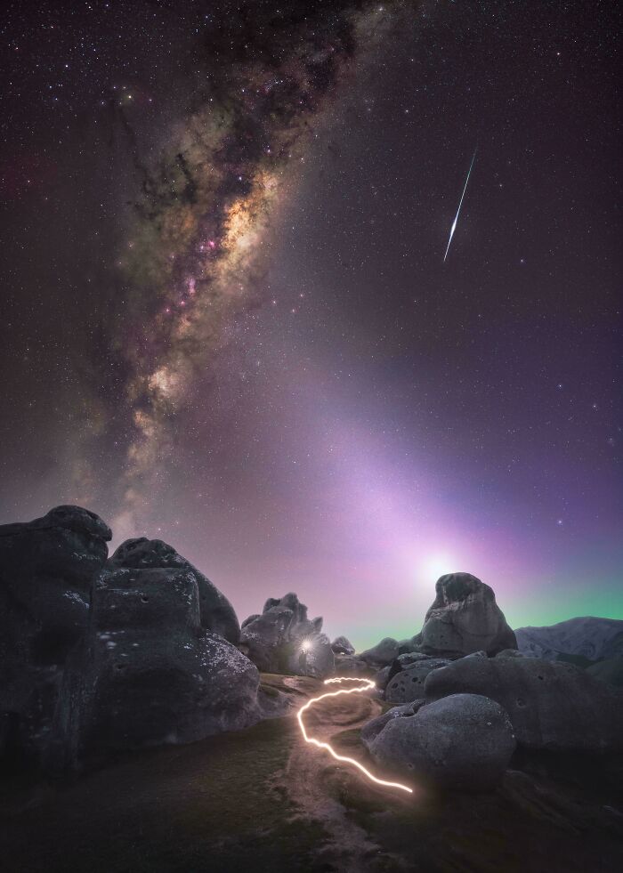 Milky Way and meteor streak over rocky landscape with glowing path, showcasing rare natural phenomena at night.