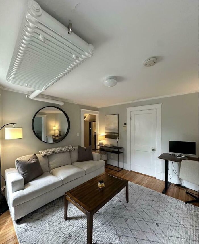 Living room with couch, round mirror, desk, and radiator installed awkwardly on the ceiling in a dumb interior design decision.