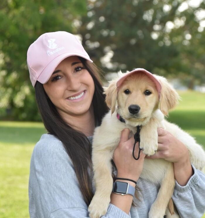 Young woman and golden retriever puppy both wearing pink hats outdoors, enjoying living their best pet summer life.