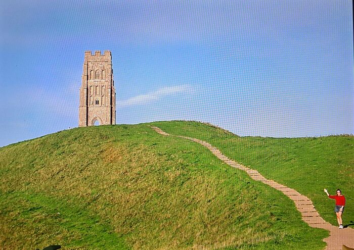 Person on a path waving near a historic tower on a grassy hill, capturing a moment for people to share found photos.