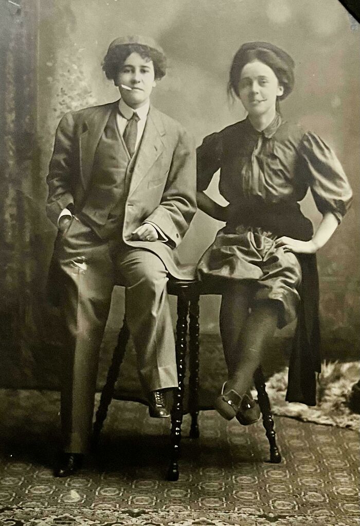 Two people share found photos in a vintage black and white portrait with formal early 20th century attire.
