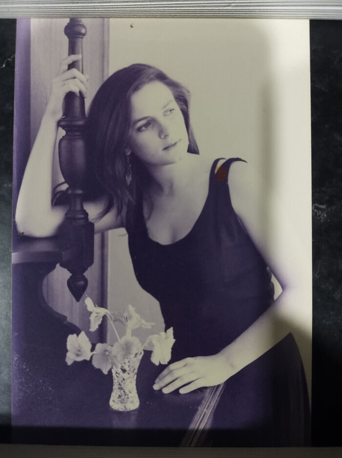 Young woman in a black dress posing thoughtfully near a vase of flowers in a vintage people-share-found photo setting.