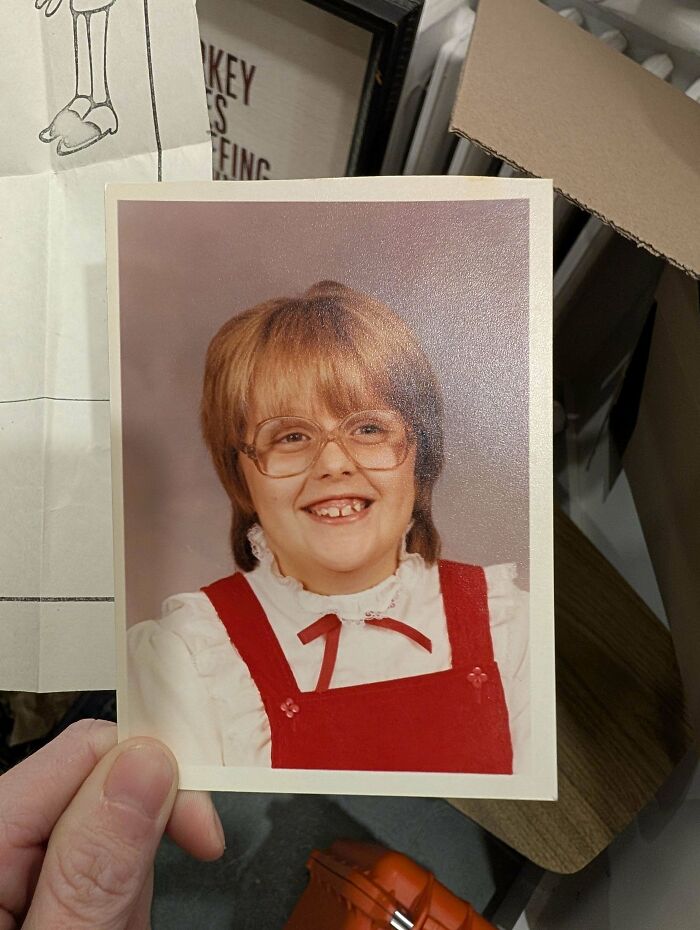 Hand holding a found photo of a smiling girl with glasses, wearing a red dress and white blouse indoors.
