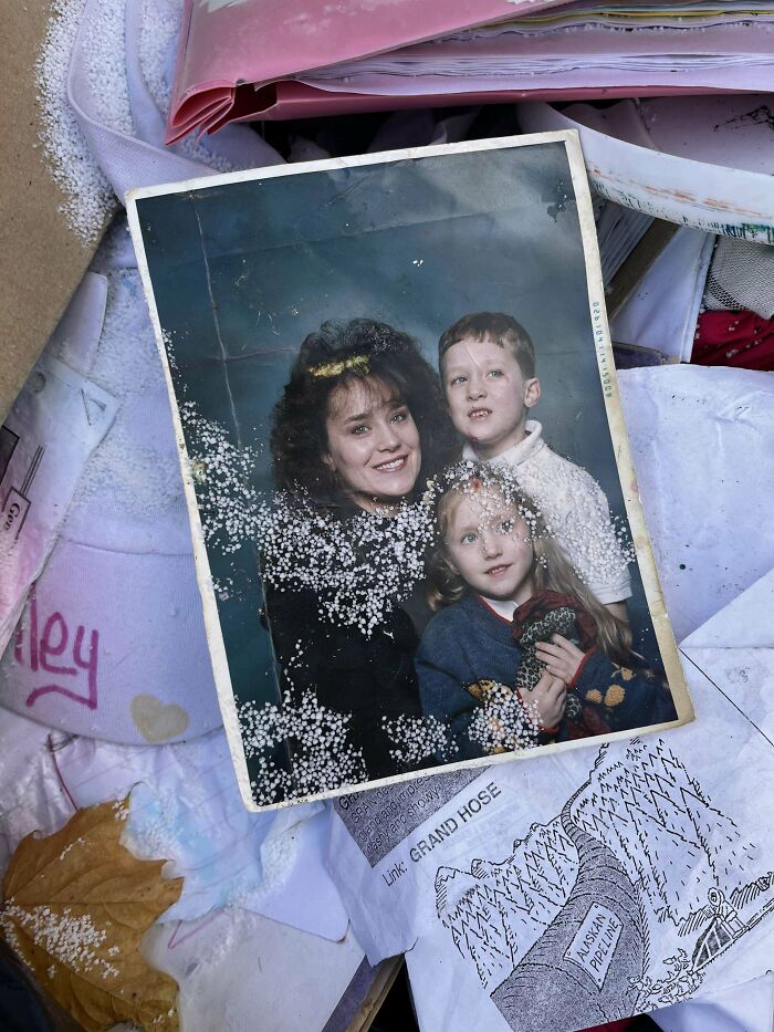 Old found photo of a smiling woman and two children, partially damaged, lying among discarded papers and fabric.