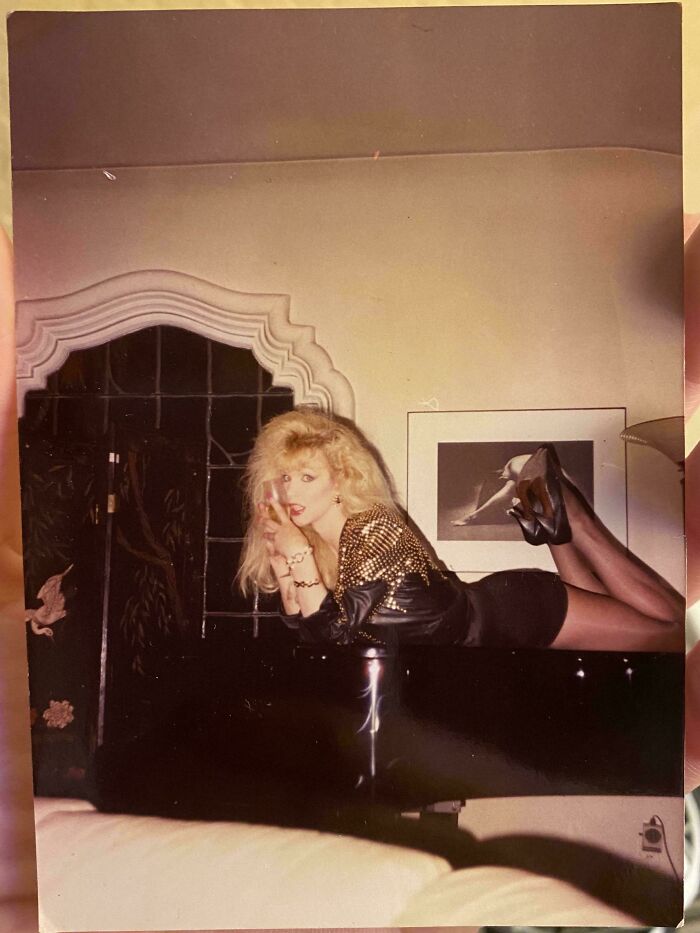 Person lying on a piano wearing a studded jacket and heels in a vintage setting, a found photo shared by people.
