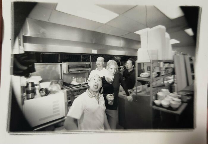 Group of people sharing found photos in a kitchen, with one person making a funny face and others posing casually.