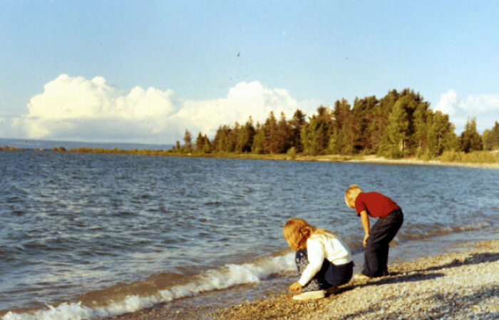 Two children on a rocky beach by the water, sharing a moment exploring found photos on a sunny day.